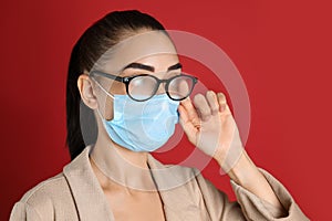 Woman wiping foggy glasses caused by wearing medical mask on red background