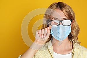 Woman wiping foggy glasses caused by wearing medical mask on yellow background. Space for text