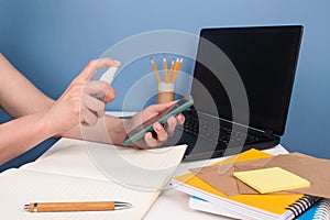 Woman wiping smartphone screen with disinfectant spray or sanitizer sitting on modern workspace. Disinfecting and cleaning