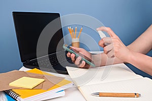 Woman wiping smartphone screen with disinfectant spray or sanitizer sitting on modern workspace. Disinfecting and cleaning
