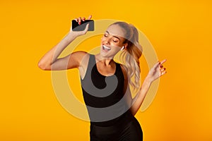 Woman In Wireless Earbuds Holding Smartphone And Dancing, Studio Shot