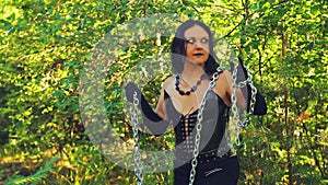 A woman of a witch in black clothes with a chain in her hands in the forest on a bright day. Halloween. Gothick style.