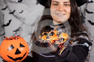 A woman in a witch costume in a hat and gloves in the background with bats holds candy and laughs. Halloween people