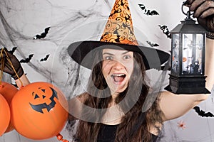 A woman in a witch costume in a hat and gloves in the background with bats holds pumpkin balls, glowing lantern and