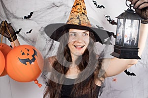 A woman in a witch costume in a hat and gloves in the background with bats holds pumpkin balls, glowing lantern and