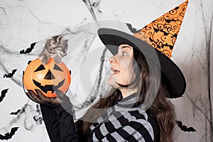 A woman in a witch costume in a hat holds a bucket pumpkin with cat. Halloween people and animal.