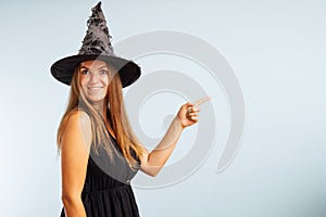 Woman in witch costume pointing with finger at copyspace side and looking at camera on blue background. Happy Halloween