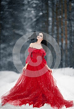 Woman witch in red dress and with raven on her shoulder in snowy