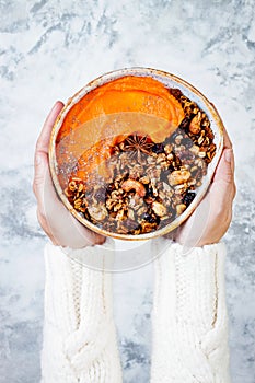 Woman in woolen sweater holding bowl with cinnamon granola, coconut yogurt, chia seeds and pumpkin puree. Healthy vegan smoothie