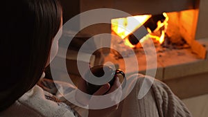 Woman in woolen sweater holding cup of hot tea and looking at burning fireplace