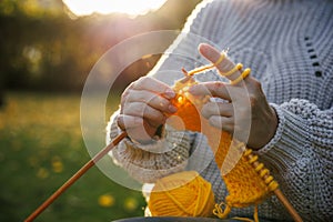 Woman in woolen sweater sitting outdoors and knits warm clothing of yellow yarn