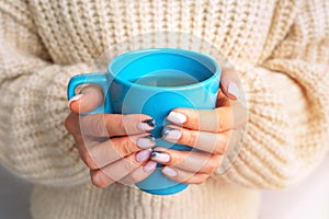 Woman in woolen sweater warming cold hands with a mug of hot tea or coffee in the morning sunlight