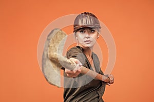 woman worker in protective helmet and boilersuit hold shovel on orange background, industry