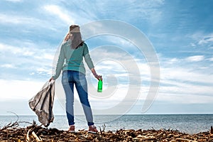 The woman worker stands on the shore of the sea. She holds a garbage bag in one hand and a dirty bottle in the other. The