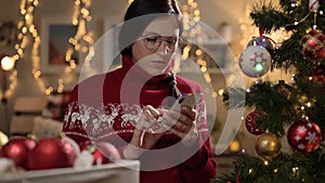 Woman working on Christmas Eve. Pensive woman decorates christmas tree and is distracted by typing message in smartphone