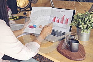 Woman working at home office desk using laptop business financial document chart and graph on wooden table with coffee cup. Woman