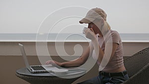 A woman is working on a laptop on the balcony of a hotel room overlooking the sea on a sunny summer day.