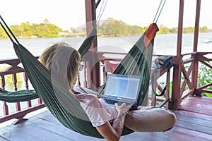 Woman working with laptop on hammock hanging in tourist resort on the Mekong River, Laos. Concept of millenials working around the photo