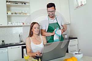 Woman working on laptop in kitchen as boyfriend prepares meal