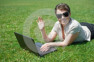 Woman working on laptop in summer outdoor, lie on your belly on green grass meadow, greeting gesture, happy people, city park