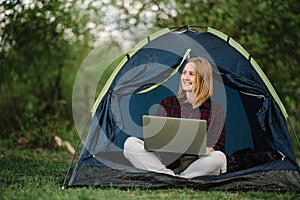Woman working on laptop in tent in nature. Young freelancer sitting in camp. Relaxing in camping site in forest, meadow. Remote