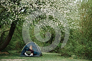 Woman working on laptop in tent in nature. Young freelancer sitting in camp. Relaxing in camping site in forest, meadow. Remote