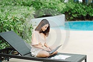 A woman working at the swimming pool with her laptop computer holding paperwork in her hand. It is imperative for young Asian