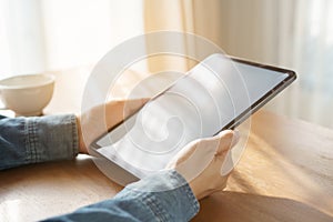 A woman working on tablet in the morning on wood table at office. The sun shines light on her hand when using tablet and it`s