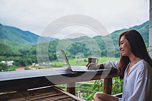 A woman working and typing on laptop computer while sitting on balcony with mountains and green nature background