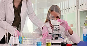 Woman works in laboratory and looks at microscope and fellow scientist puts test tubes with liquids