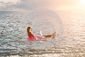 Woman works on laptop in sea. Freelancer, blond woman in sunglases floating on an inflatable big pink donut with a