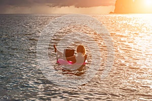 Woman works on laptop in sea. Freelancer, blond woman in sunglases floating on an inflatable big pink donut with a photo