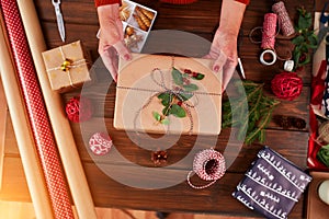 Woman wrapping gift box with decorating items on wood table, close up, top view.