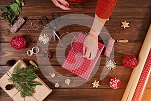 Woman wrapping gift box with decorating items on wood table, close up, top view.