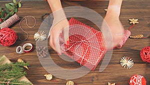 Woman wrapping gift box with equipment and decorating items on wood table, preparing for celebrating Christmas holidays