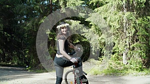 Woman in wreath of daisies with bike alone in the bright green forest looking at the camera and greeting the spectator