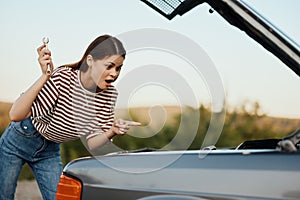 A woman with a wrench with a smile looks happily under the open hood of her car and repairs it from a roadside breakdown
