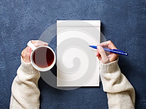 woman writes in notebook on dark blue table, hand in shirt holding a pencil, cup of tea, sketchbook drawing, top view