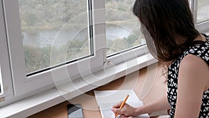 Woman is writing a letter in notebook sitiing on the balcony at home. View of the river. The rain outside the window.