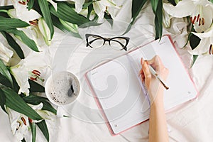 Woman writing in notebook while lying in the white bedroom. Good morning and weekend concept in flat lay style