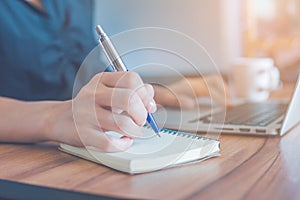 Woman is writing on a notebook with a pen and using a laptop to work in the office