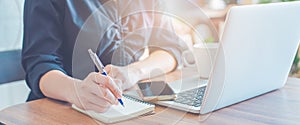 Woman is writing on a notebook with a pen and using a laptop to work in the office