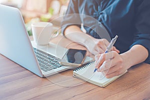 Woman is writing on a notebook with a pen and using a laptop to work in the office