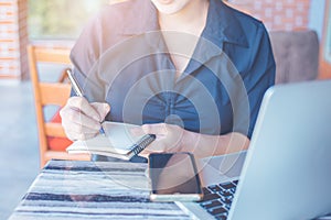 Woman is writing on a notebook with a pen and she is using a mobile phone and computer laptop in the office