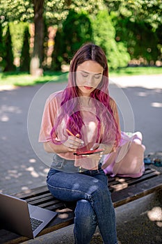 Woman writing in a notebook sitting on a wooden bench in the park. Girl working outdoors on portable computer, copy