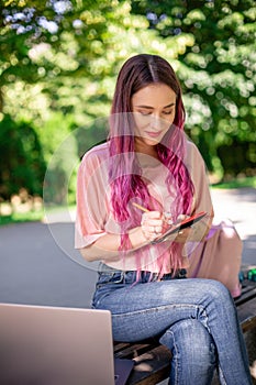 Woman writing in a notebook sitting on a wooden bench in the park. Girl working outdoors on portable computer, copy