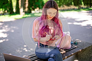 Woman writing in a notebook sitting on a wooden bench in the park. Girl working outdoors on portable computer, copy