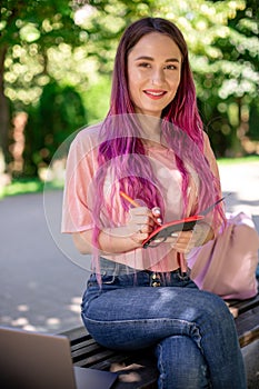 Woman writing in a notebook sitting on a wooden bench in the park. Girl working outdoors on portable computer, copy