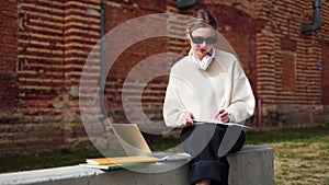 Woman writing in notepad while looking at laptop screen and resting outdoors