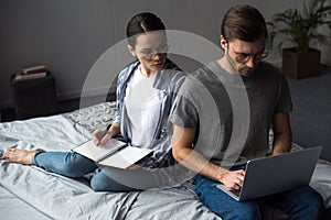 Woman writing in notepad and looking at man working on laptop while sitting